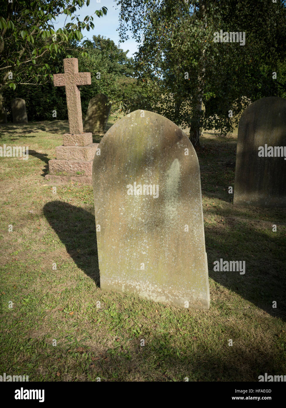Plain lapide con parte superiore arcuata oscurato dalla presenza di foglie contro lo sfondo di colore verde in un cimitero con headstone cross in background Foto Stock