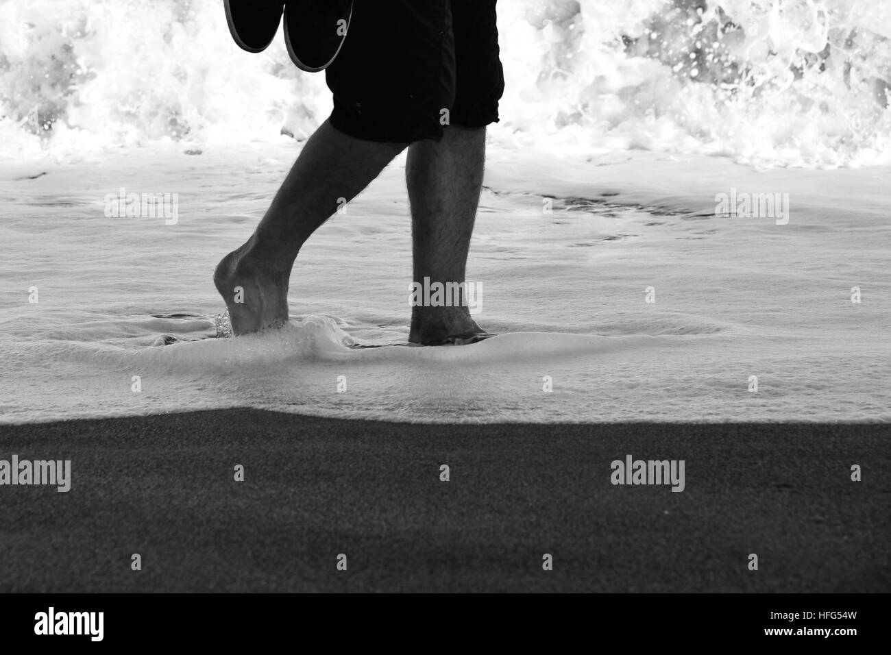 Immagine in bianco e nero di un uomo che trasportava formatori passeggiando attraverso il surf in bordo di mare Foto Stock