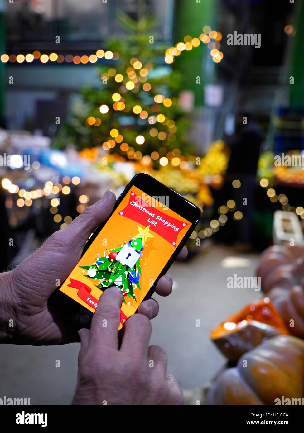 Maschio maturo mani, tenendo smartphone iPhone 7 Plus, con protetti da pin a tema natale shopping list app sulla schermata interattiva. Atmosferica stagionale albero di Natale luci e Mercato di Borough si spegne dietro London REGNO UNITO Foto Stock