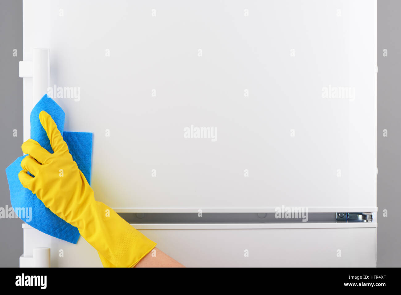 Mano di donna gomma gialla di guanto protettivo pulizia frigorifero bianco con rag di colore blu su sfondo grigio Foto Stock
