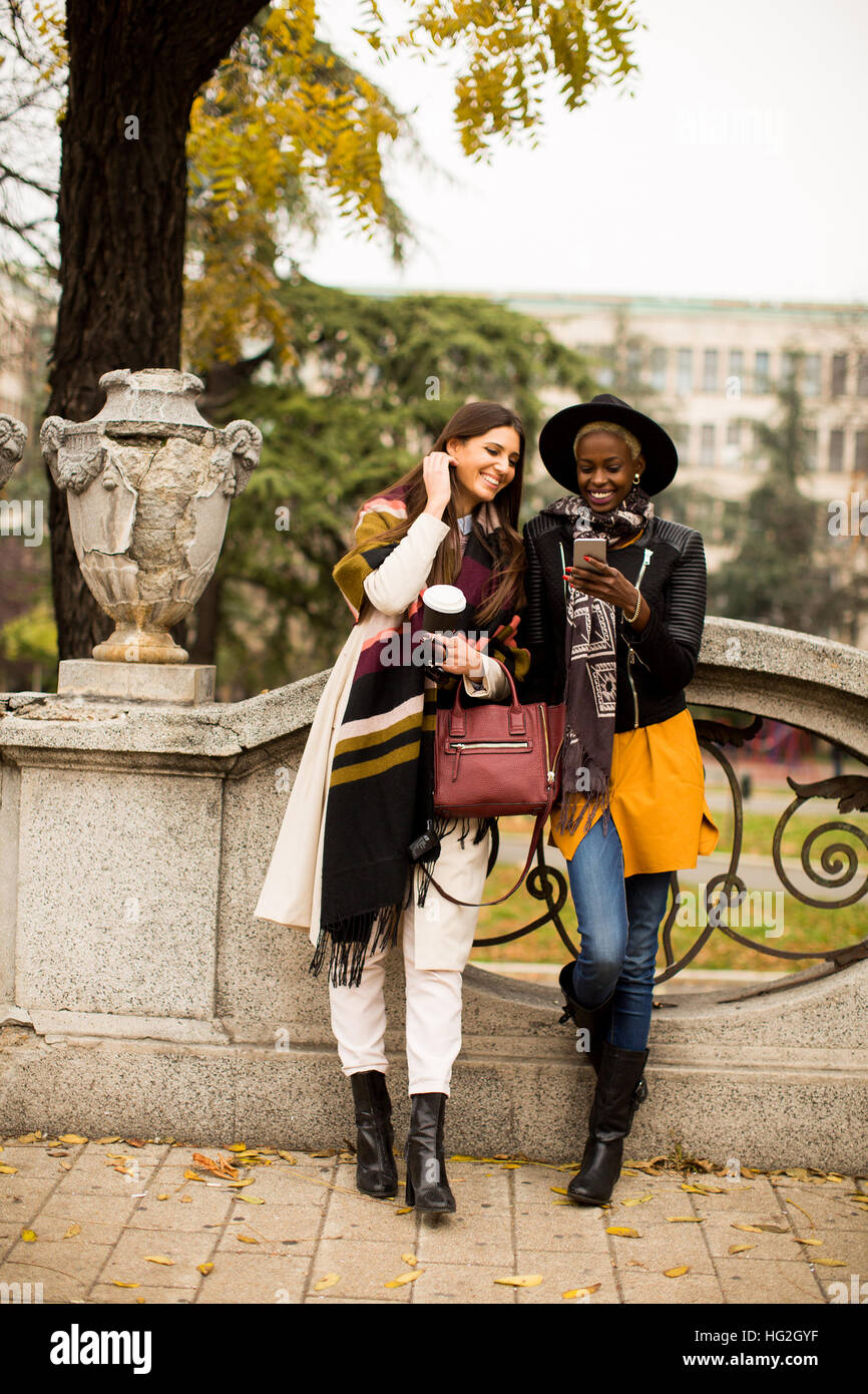 Americano africano e caucasici che pongono la donna fuori con il telefono cellulare e una tazza di caffè per andare in autunno Foto Stock