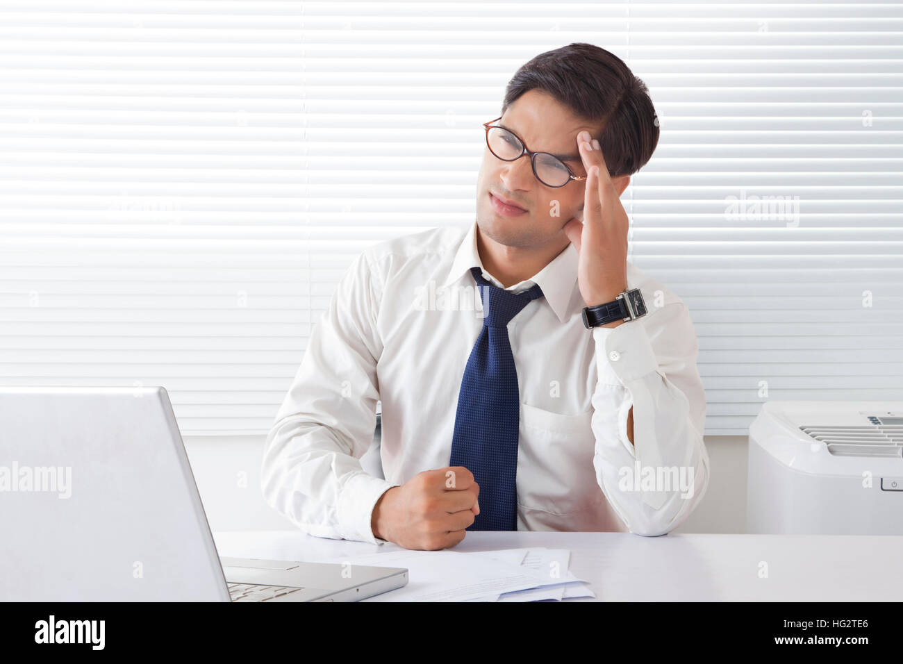 Giovane uomo cercando frustrati e arrabbiati tenendo la fronte seduto alla scrivania in ufficio Foto Stock