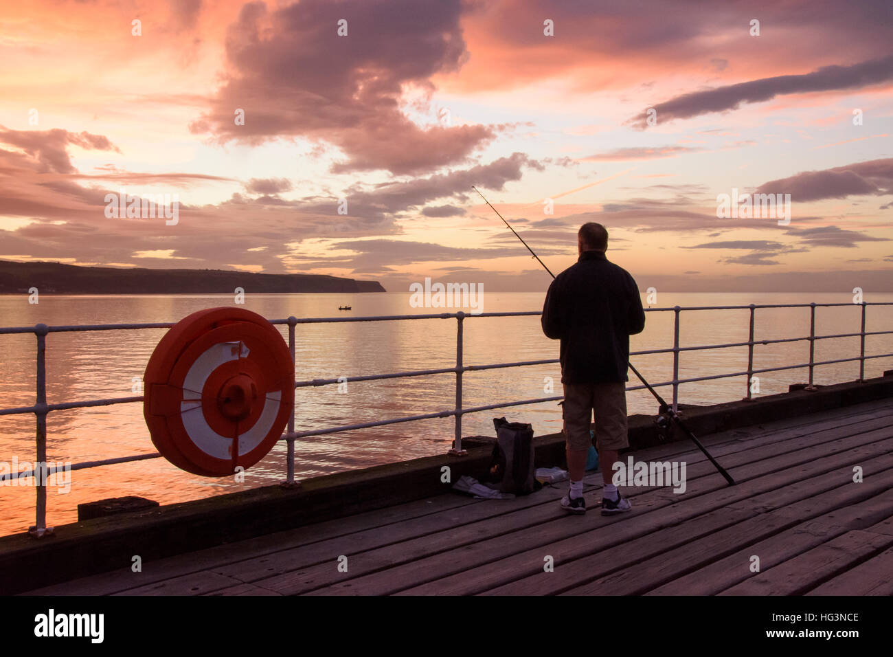 Scenic Whitby & stagliano drammatico, luminoso colorato tramonto cielo, uomo (pescatore) è la pesca in mare dal Molo Ovest - Nord Yorkshire, Inghilterra, Regno Unito. Foto Stock