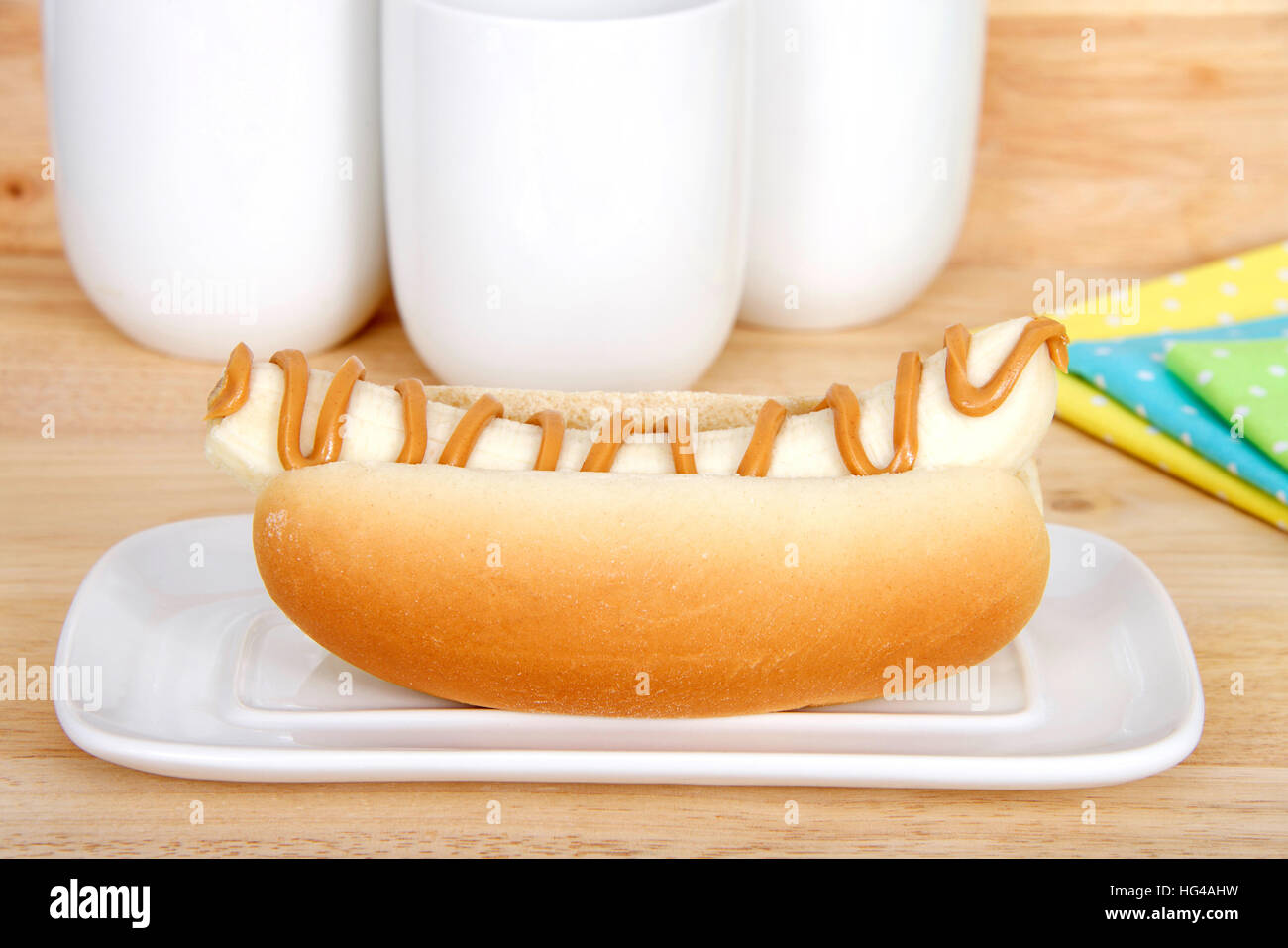 Banana con burro di arachidi in un hot dog bun servita su un bianco piastra rettangolare, giallo, blu, verde polka dot tovaglioli e bicchieri in background su legno t Foto Stock