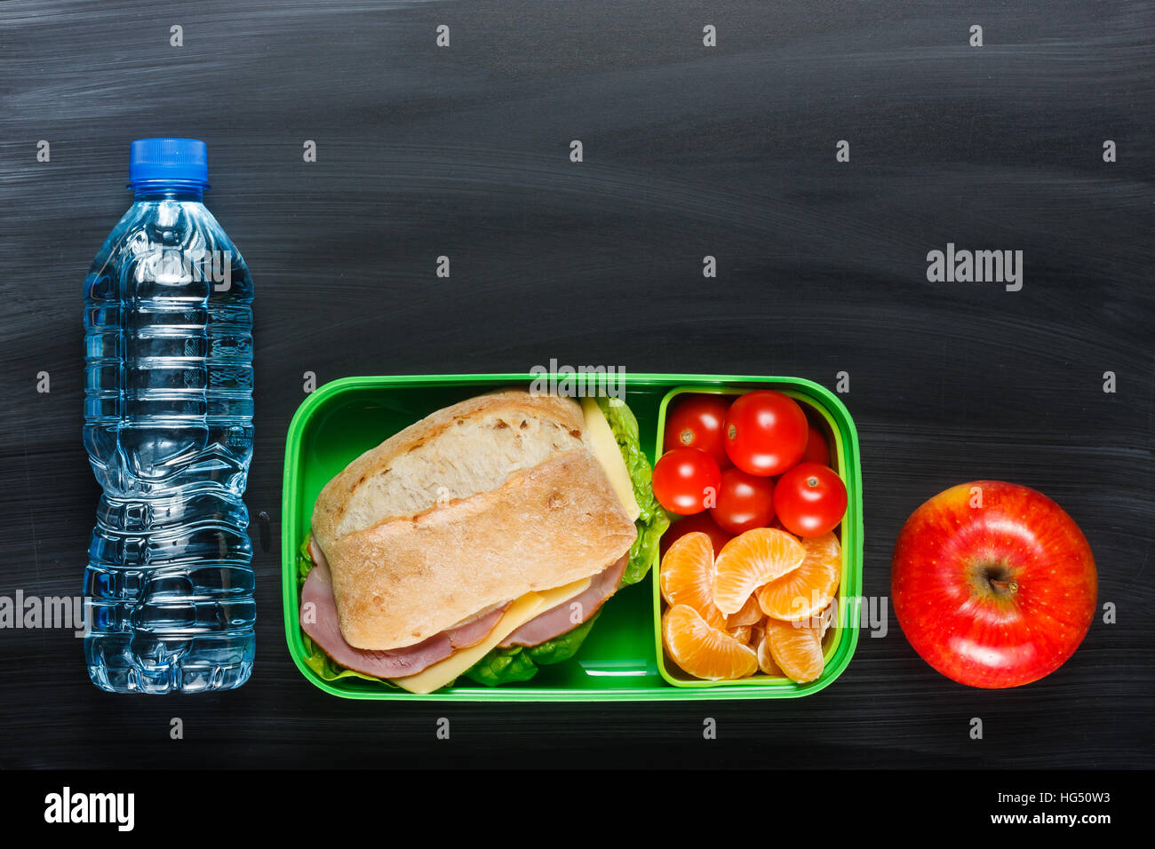 Sandwich, pomodorini, mandarino, Apple in plastica scatola di pranzo e una bottiglia di acqua sulla lavagna. Foto Stock
