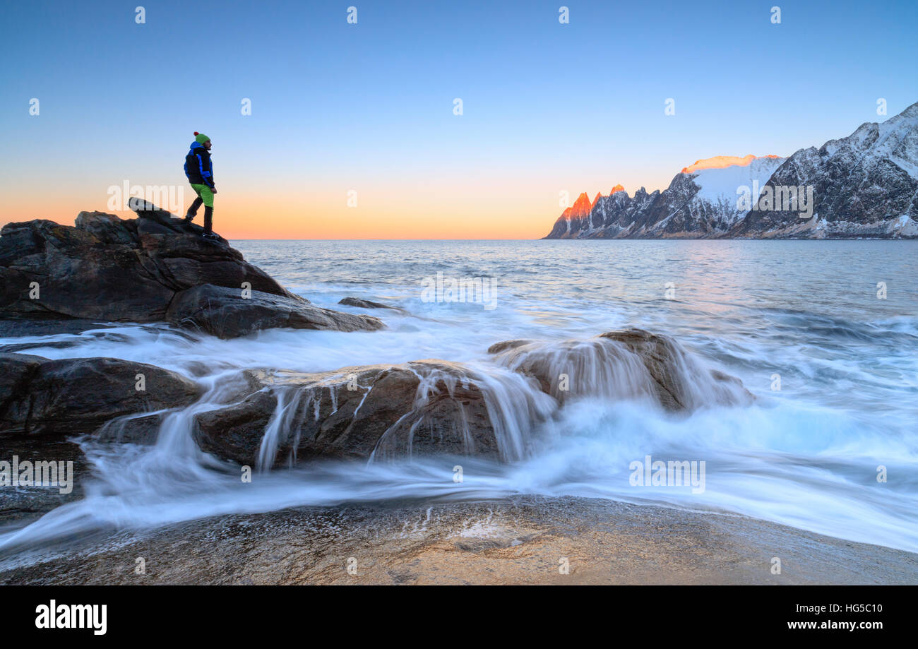 Escursionista ammira le onde del mare ghiacciato che si infrangono sulle scogliere rocciose all'alba, Tungeneset, Senja, Troms County, artiche, Norvegia Foto Stock