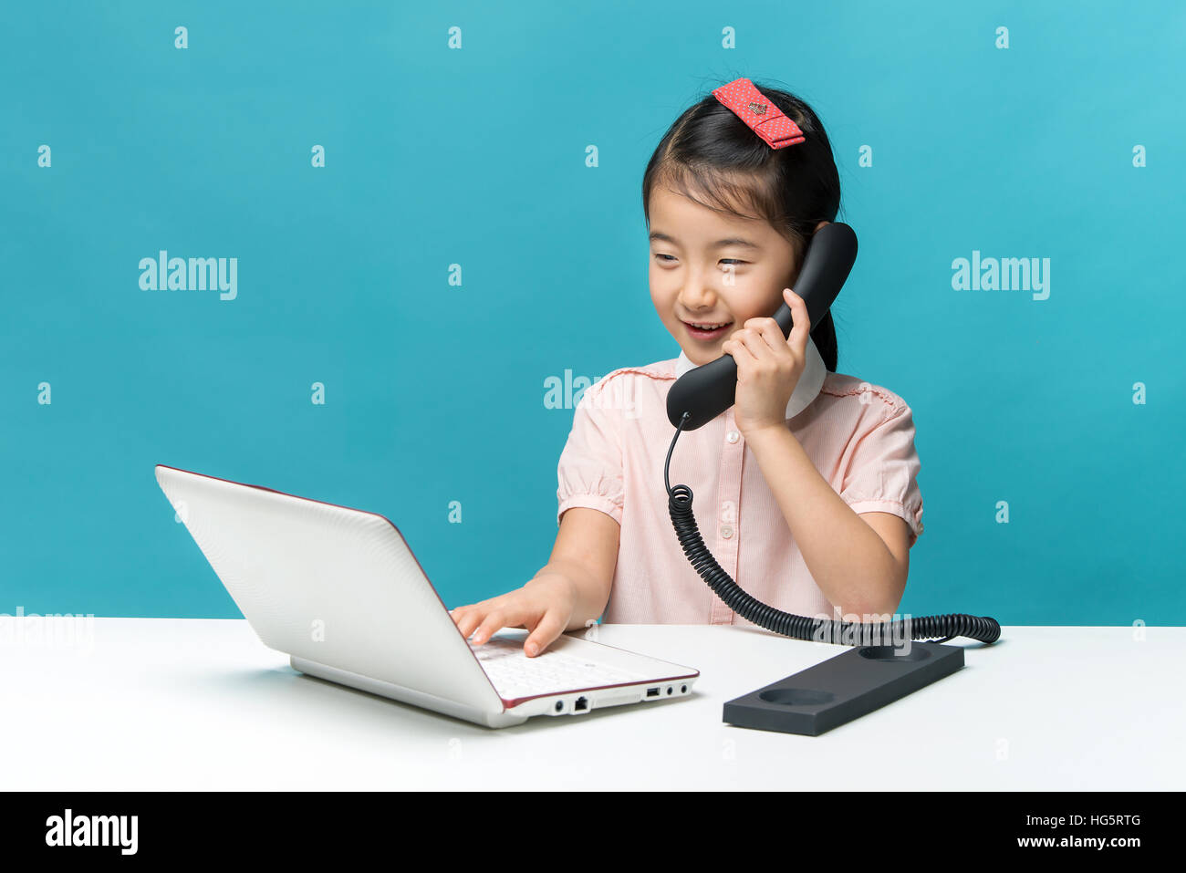 Carino asia bambina è seduta a tavola con il suo bianco portatile e un telefono, isolate su blu Foto Stock