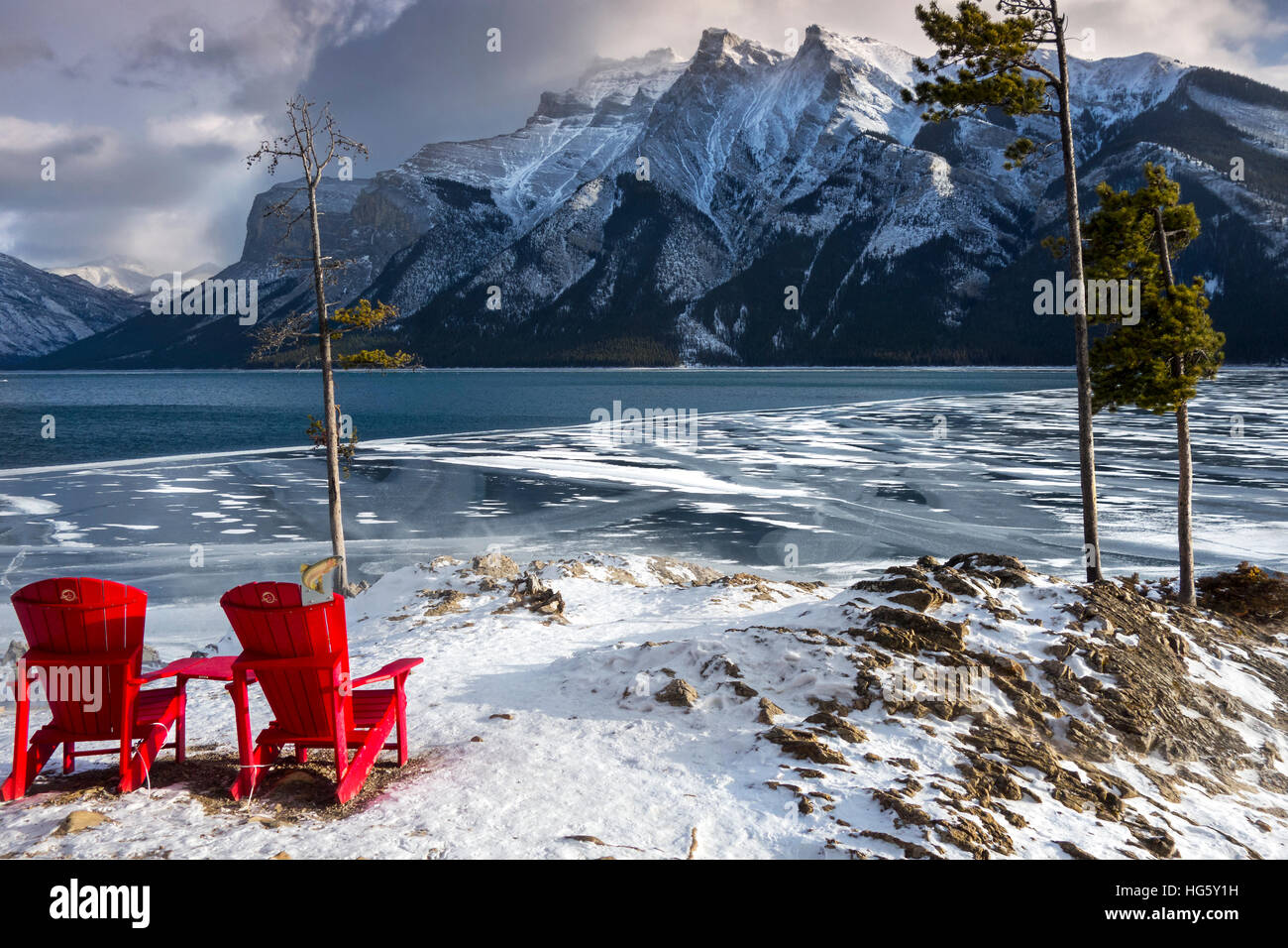 Sedie rosse Adirondack, paesaggio invernale lontano da Snowy Mountain Peak. Melting Lake Minnewanka Ice, Banff National Park, Rocky Mountains Alberta Canada Foto Stock