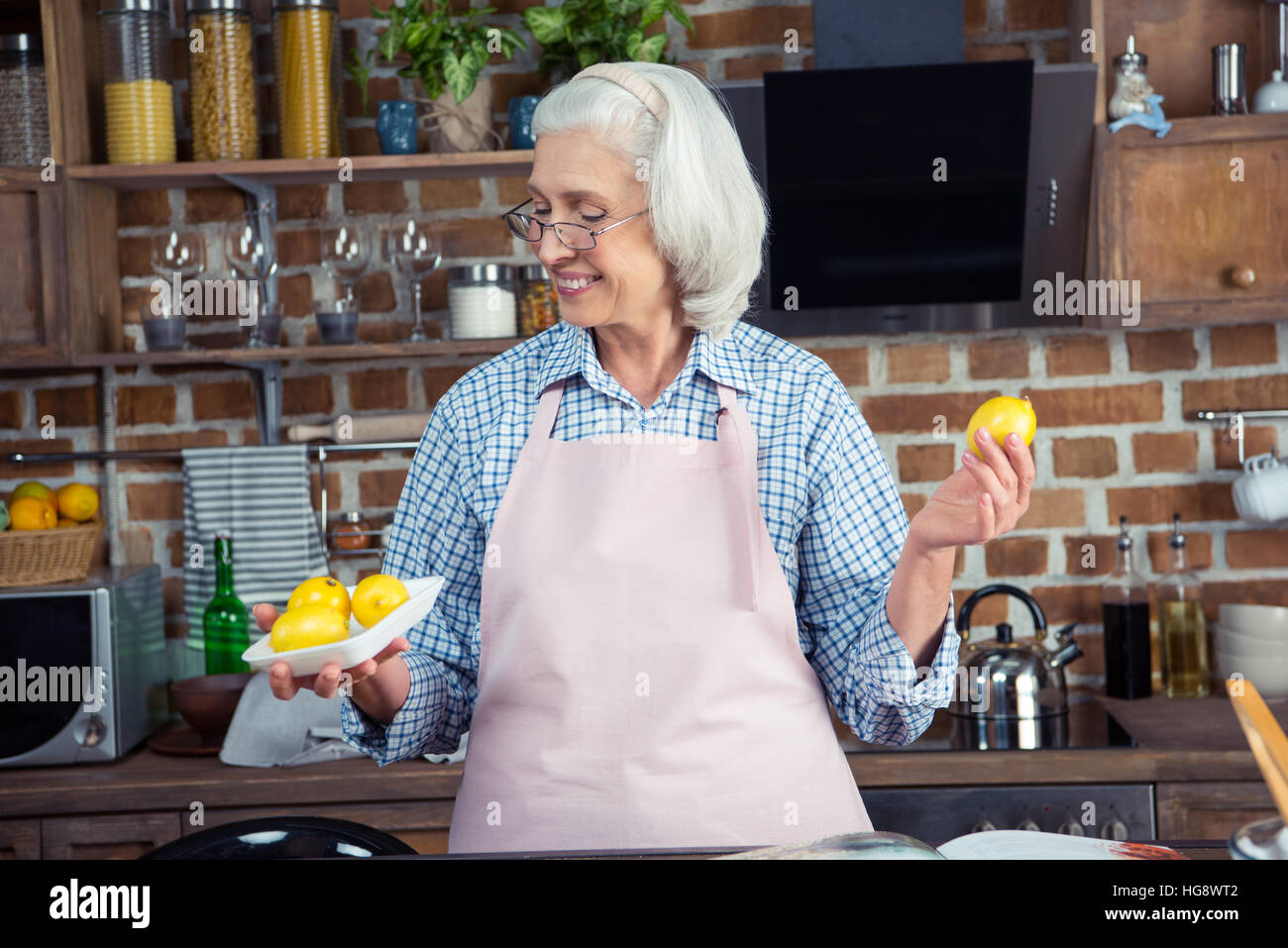 Sorridente donna senior in occhiali e grembiule guardando i limoni in cucina Foto Stock
