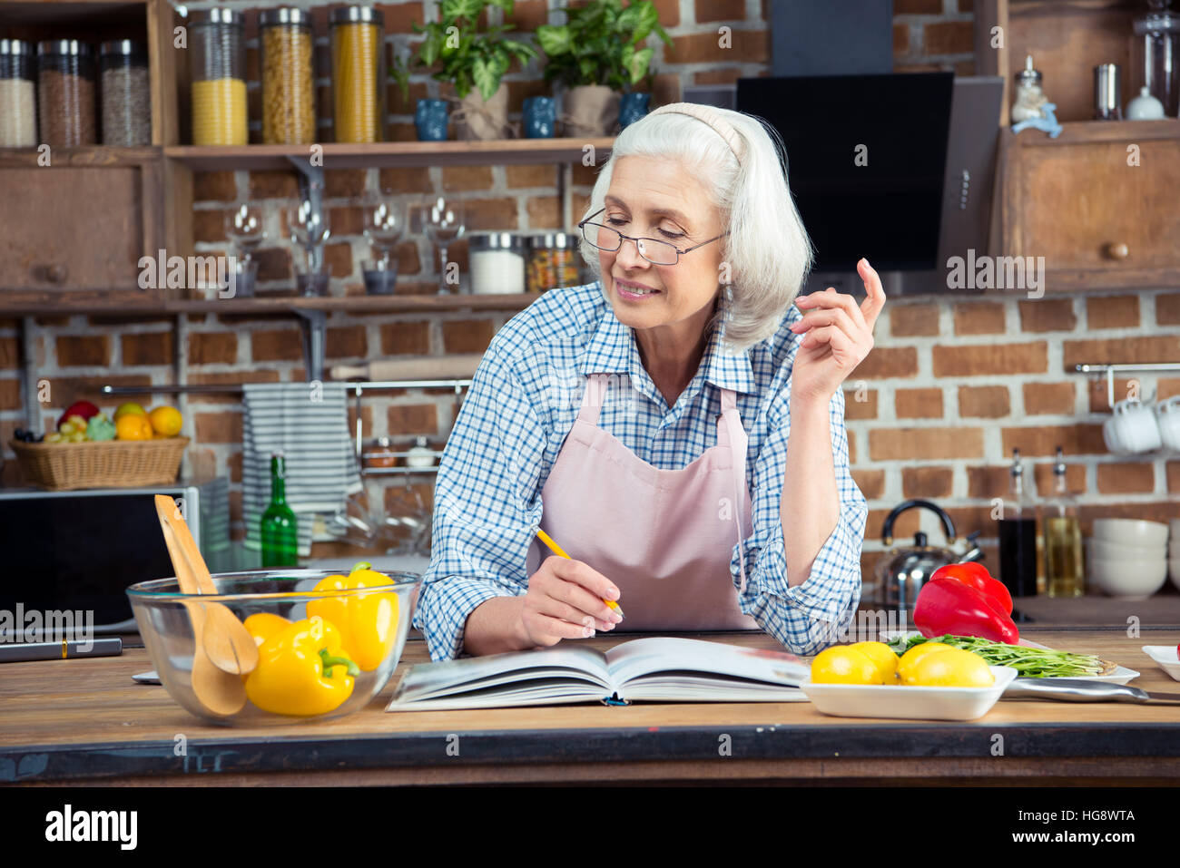 Sorridente donna senior iscritto nel libro di cucina con matita e guardando gli ingredienti Foto Stock