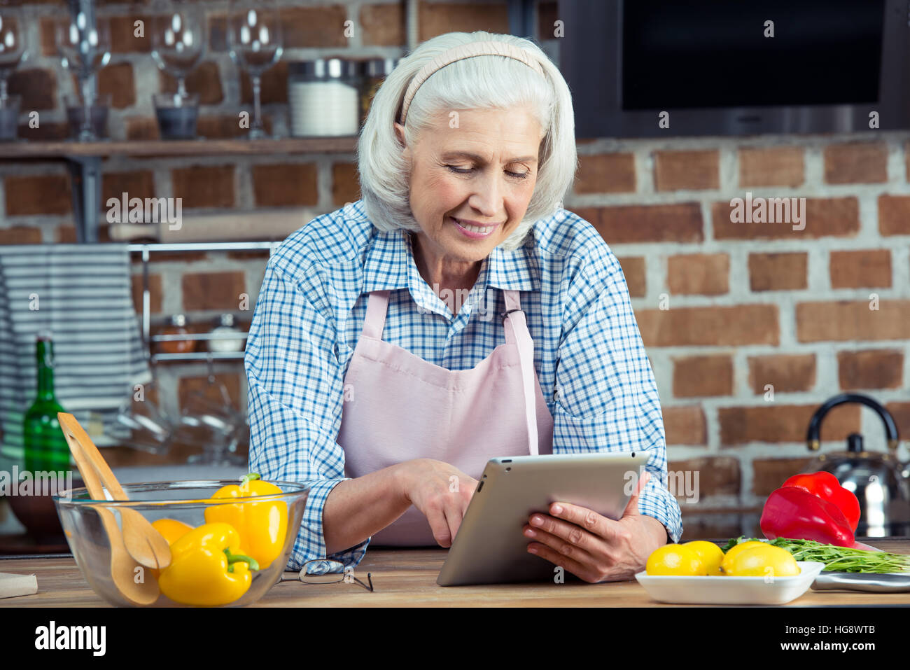 Senior sorridente donna in grembiule utilizzando digitale compressa in cucina Foto Stock