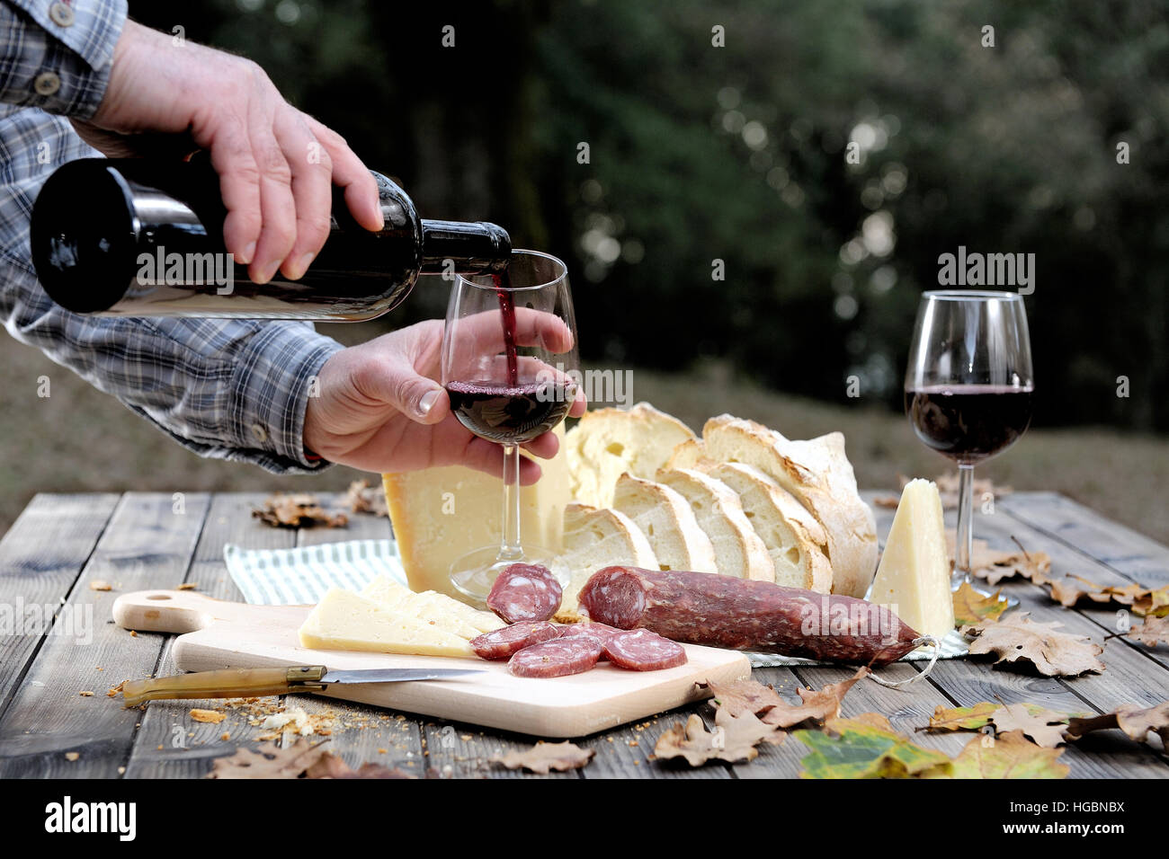 Le mani che versano il vino nel calice mentre mangia in campagna con pane, formaggio e salsicce. Foto Stock