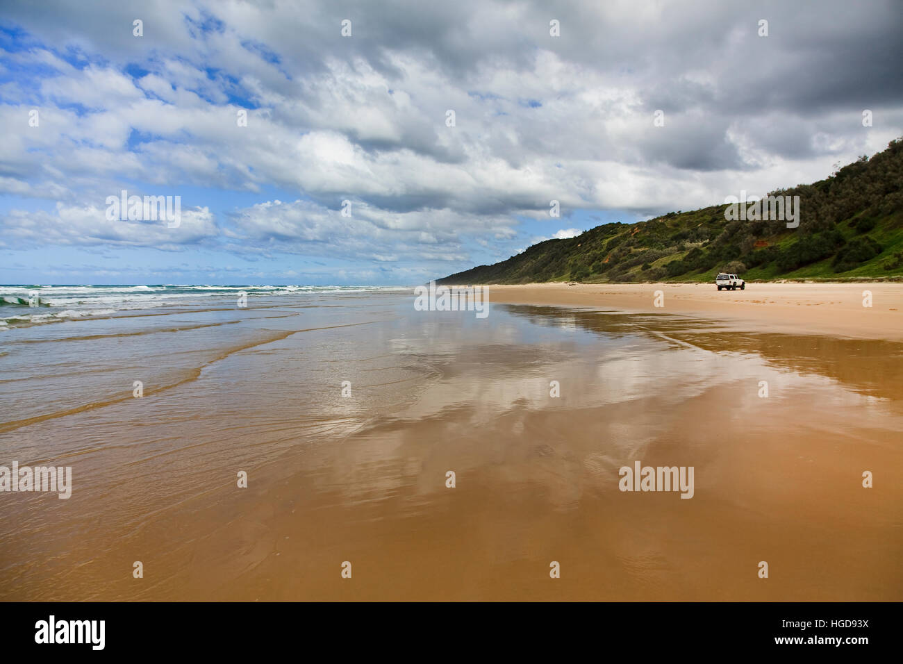 Ampio appartamento wet spiaggia sabbiosa di Fraser Island - autostrada eccellente per 4WD auto turismo. Le onde di marea riflettono le nuvole e il verde delle colline di isola di sabbia. Foto Stock