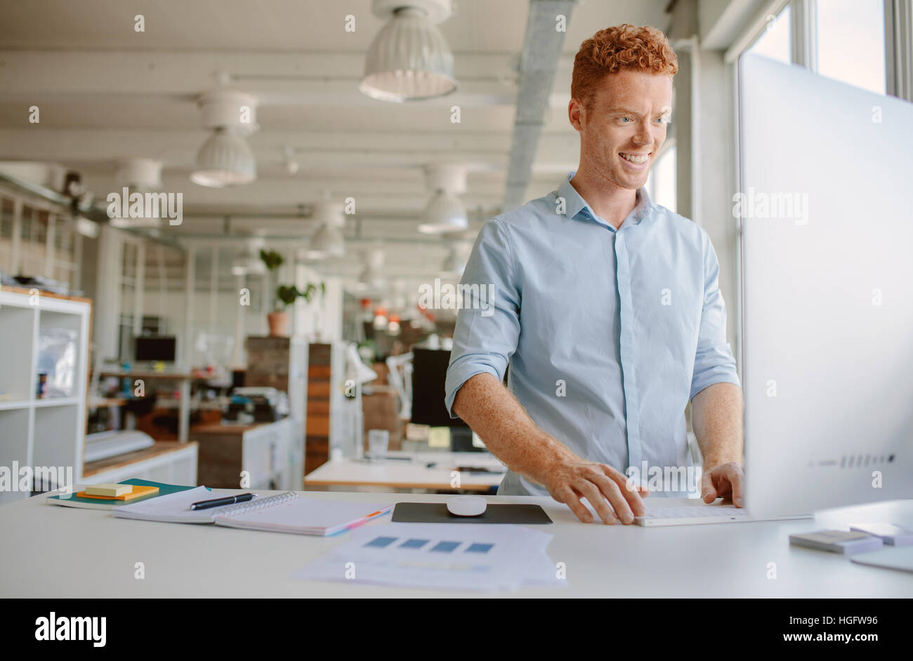 Colpo di felice giovane uomo in piedi alla sua scrivania e lavorare sul computer. Imprenditore lavora in un ufficio moderno. Foto Stock
