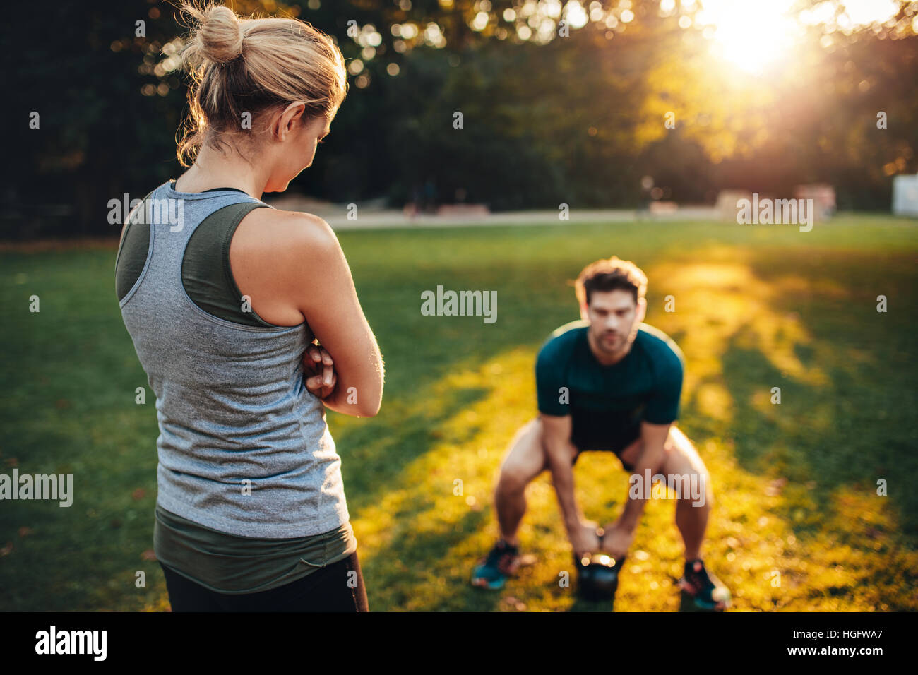 Trainer femmina in piedi nel parco con un giovane uomo che fa del peso della formazione con kettlebell in background. Foto Stock