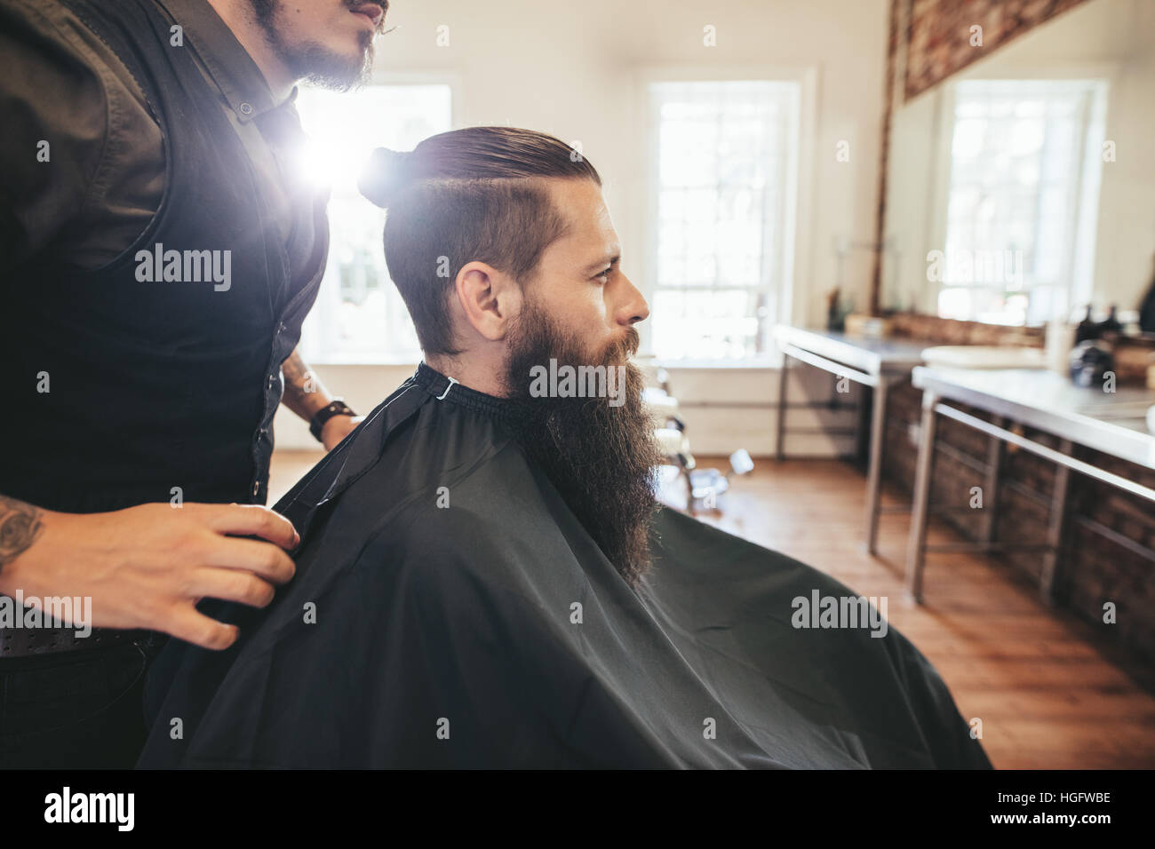Vista laterale colpo di bello uomo barbuto in un nero il taglio di capelli cape in Barberia, con parrucchiere in attesa. Uomo seduto al salone di parrucchiere. Foto Stock