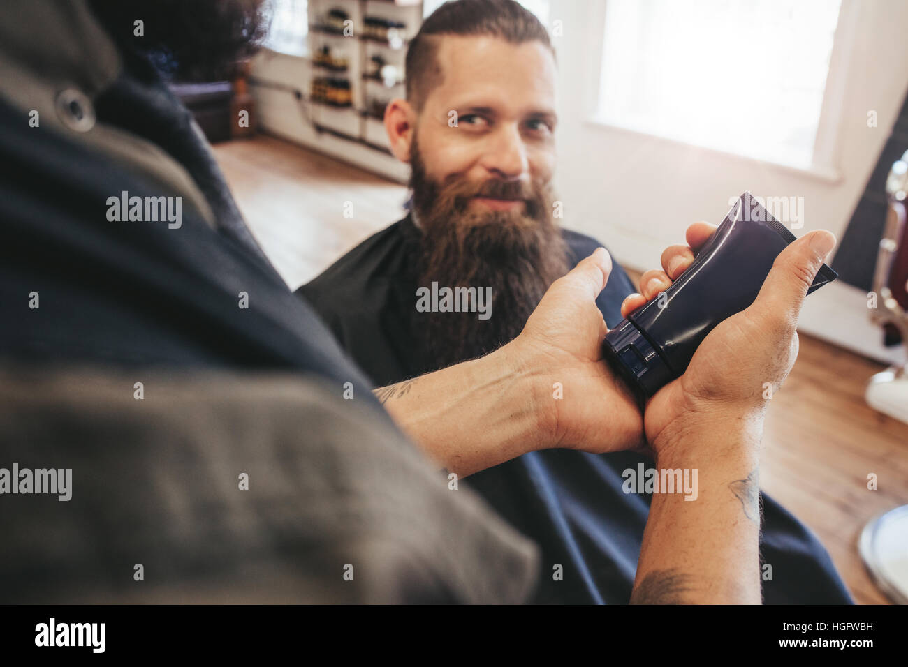 Barbiere di servire il suo cliente. Barba uomo seduto in poltrona e guardando il parrucchiere. Focus su Azienda barbiere crema cosmetica in mano. Foto Stock