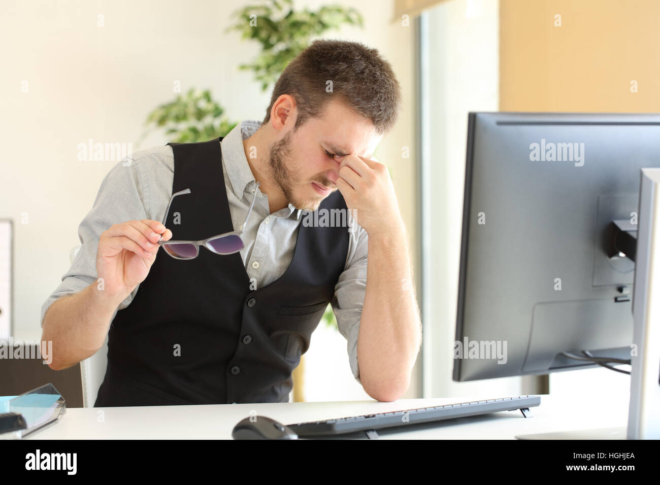 Imprenditore sofferenza affaticamento degli occhi e tenendo gli occhiali seduto in un desktop in ufficio Foto Stock