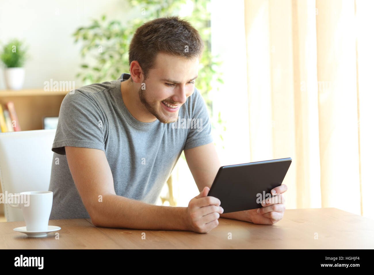 Happy guy la visione di contenuti multimediali in linea in un tablet seduto su una sedia in una tabella a casa Foto Stock