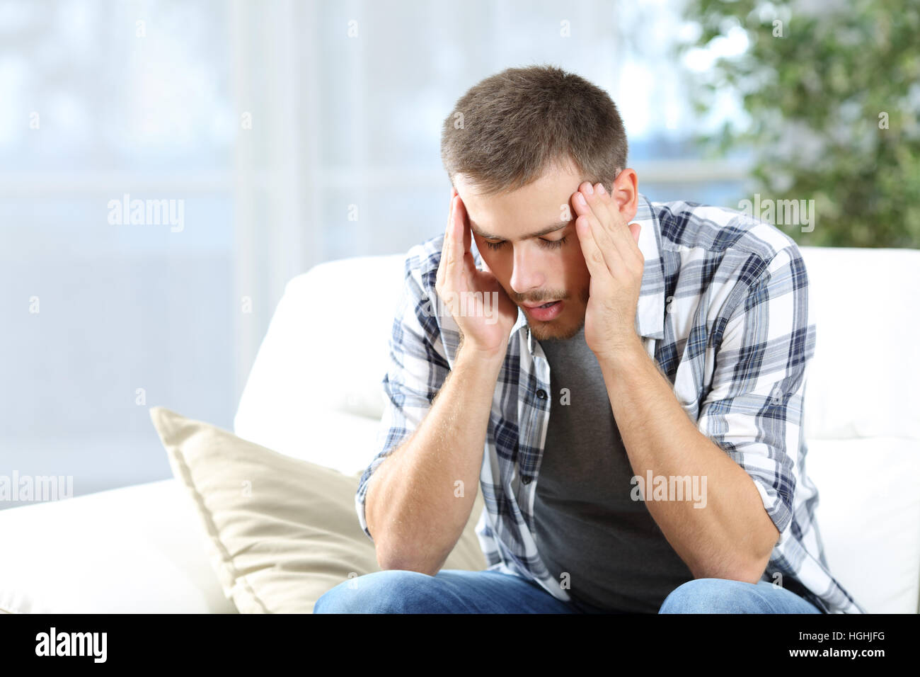 Uomo che soffre mal di testa seduto su un divano nel soggiorno di casa Foto Stock