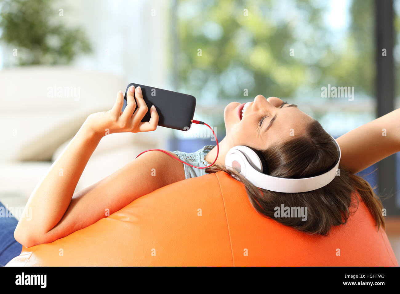 Teen ascolto di musica con cuffie e smart phone giacente su un pouf arancione nel salotto di casa Foto Stock