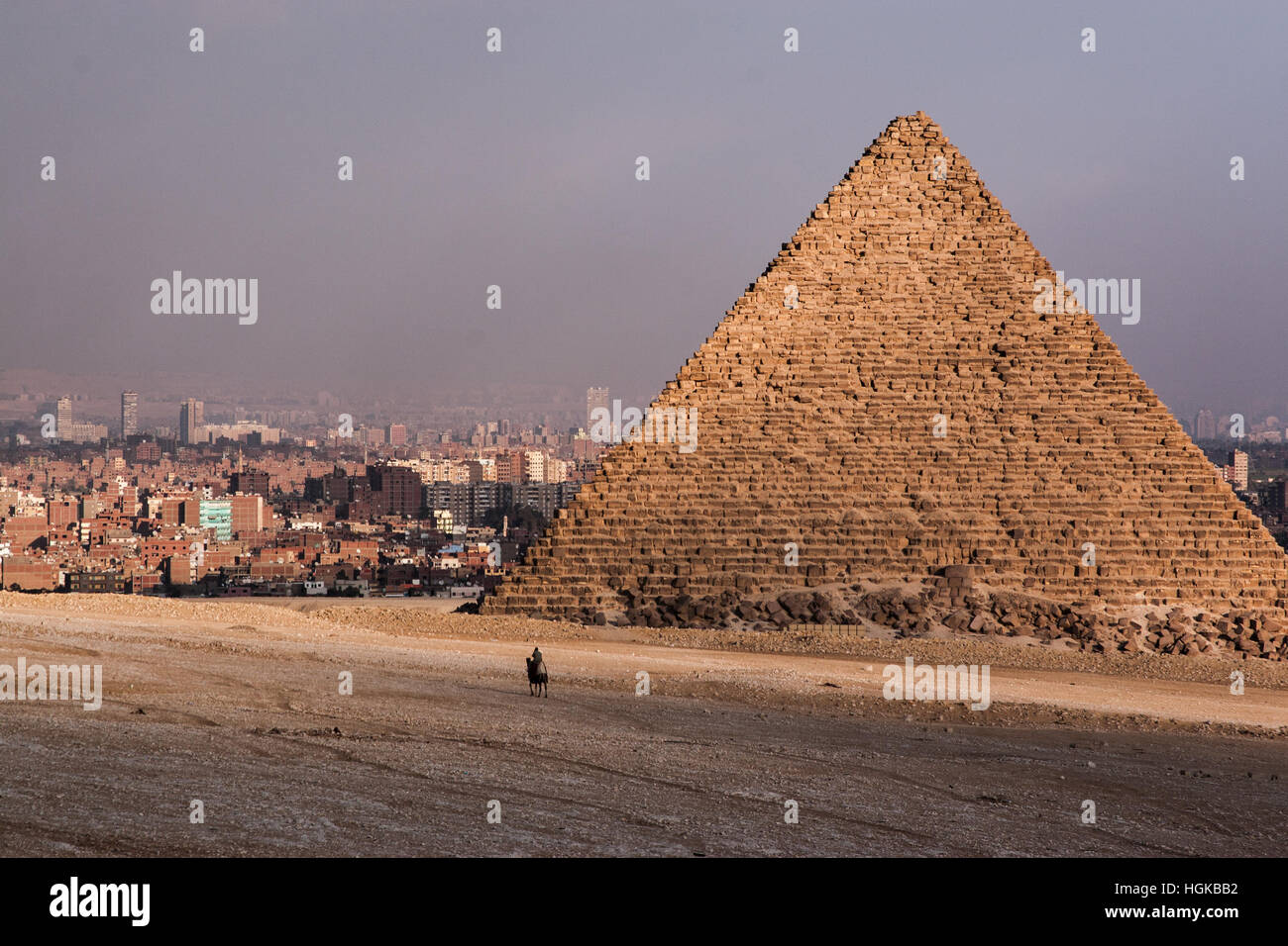 Lone rider su un cammello vicino alla base di una delle piramidi di Giza con la città del Cairo in background. Foto Stock