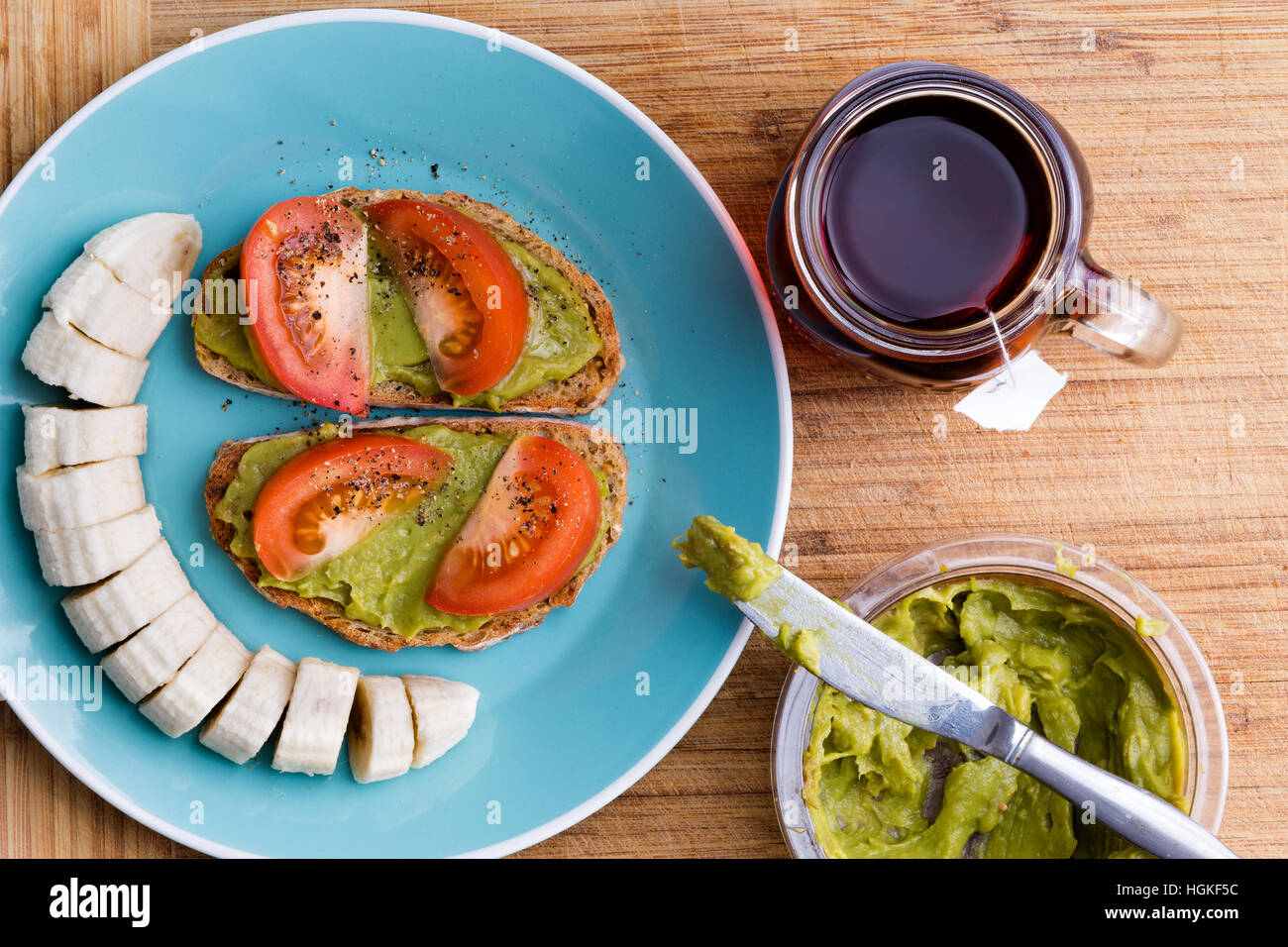 Colazione energetica con il grano intero pane di grano condito con chunky guacamole e pomodori, servita su piastra blu con banane a fettine e tè caldo sul Foto Stock