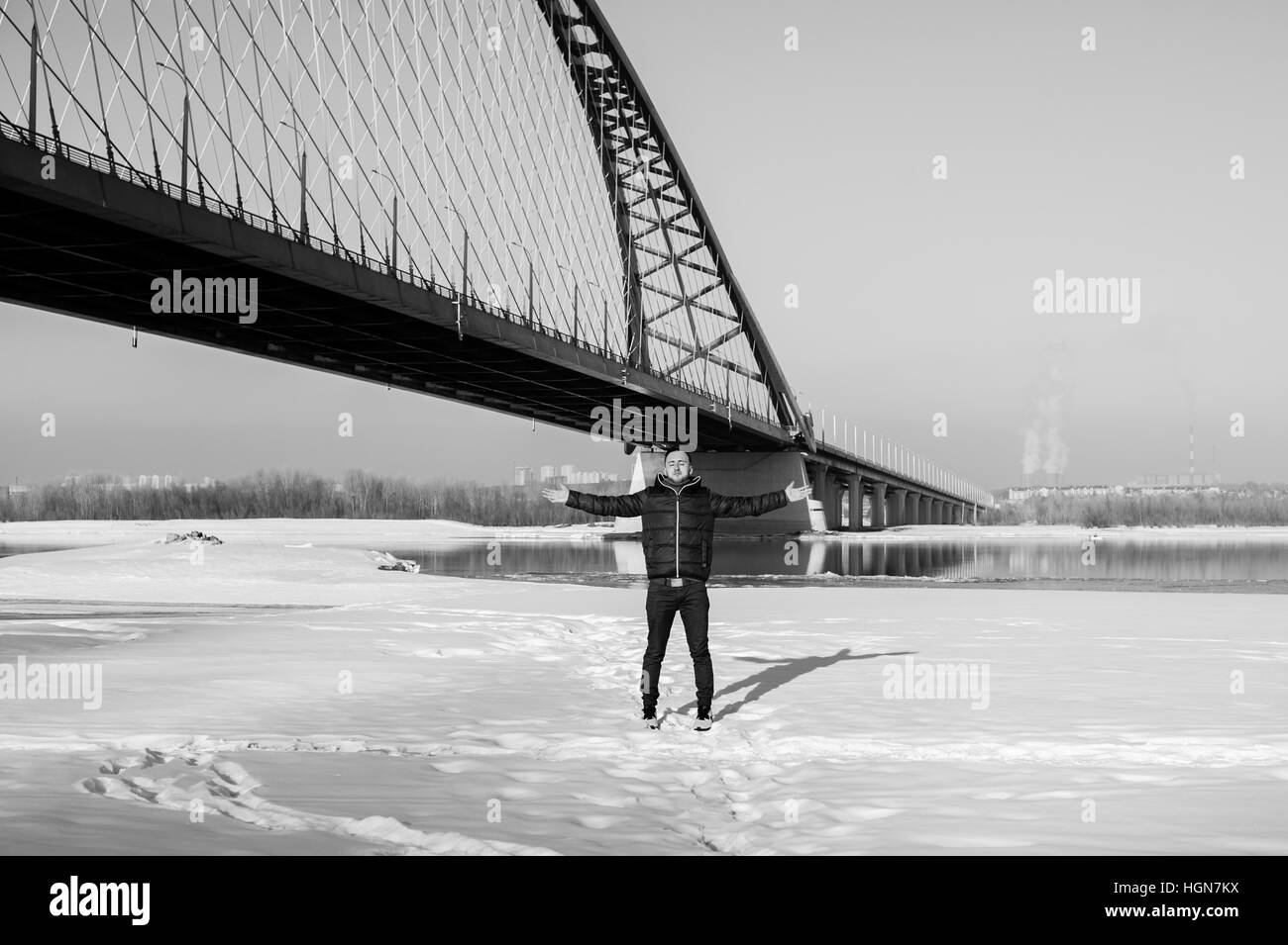 Uomo in vestiti caldi con la barba sta camminando per la strada in inverno in un giorno caldo e soleggiato presso il ponte sul fiume in background Foto Stock