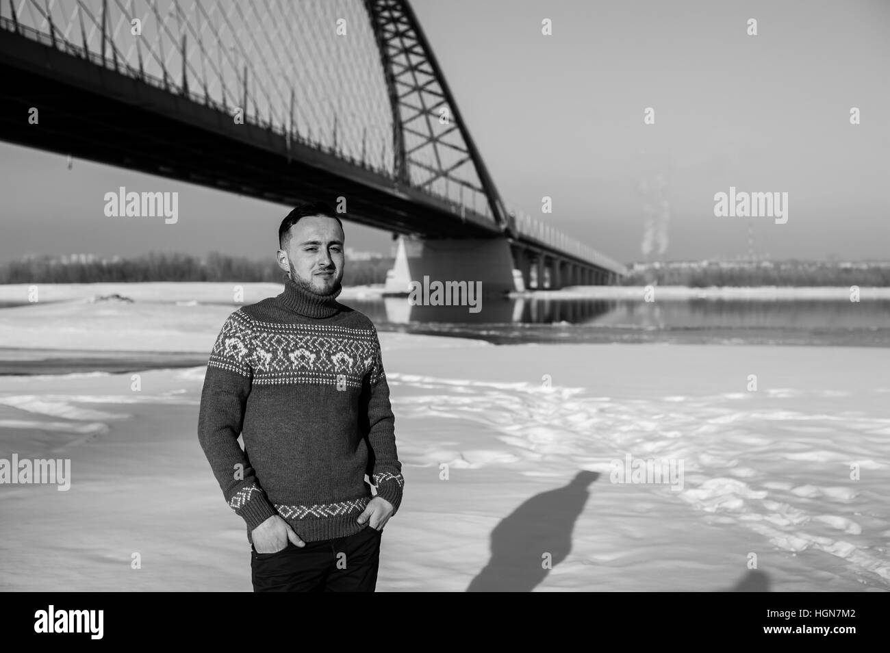 Uomo in vestiti caldi con la barba sta camminando per la strada in inverno in un giorno caldo e soleggiato presso il ponte sul fiume in background Foto Stock