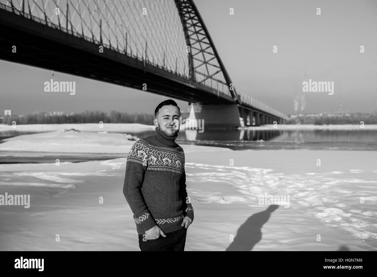 Uomo in vestiti caldi con la barba sta camminando per la strada in inverno in un giorno caldo e soleggiato presso il ponte sul fiume in background Foto Stock