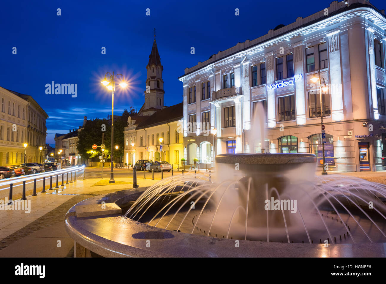 Vilnius, Lituania - 8 Luglio 2016: Grigio Fontana in marmo con getti d'acqua su accesa Rotuses Square. Il campanile della chiesa di San Nicola dietro in estate Foto Stock