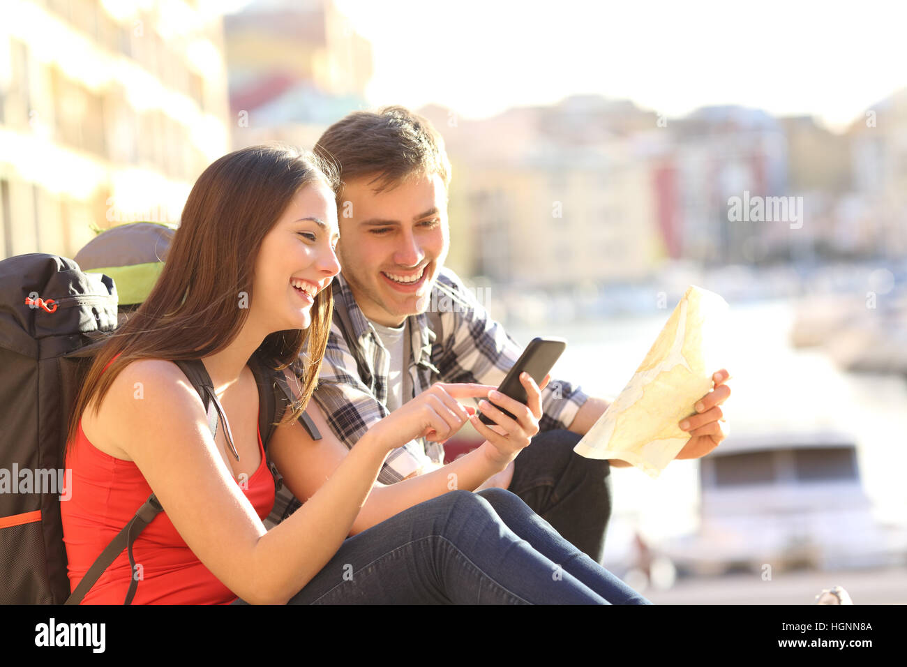 Due turisti alla ricerca posizione su uno smart phone seduto sul pavimento di una destinazione di viaggio nella costa con una calda luce in background Foto Stock