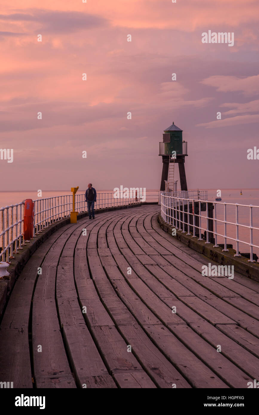 Uomo che cammina da sola a Whitby West Pier estensione, sotto un drammatico, Vivid, rosa, rosso, il cielo al tramonto - North Yorkshire, Inghilterra. Foto Stock