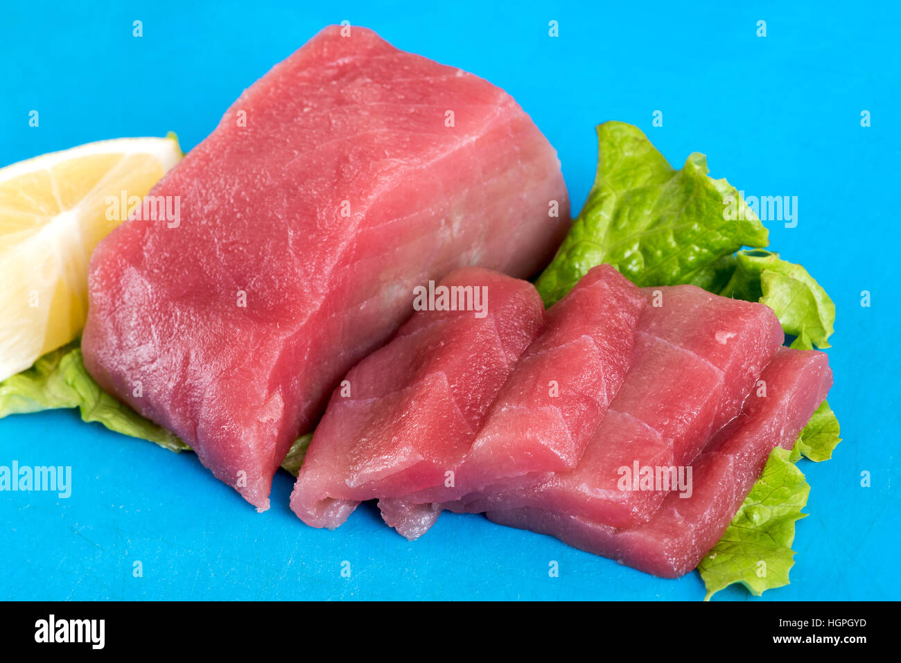 Affettato crudo fresco di filetto di tonno su una foglia di lattuga con un cuneo di limone da utilizzare per realizzare il sushi o il sashimi, colorato sfondo blu Foto Stock