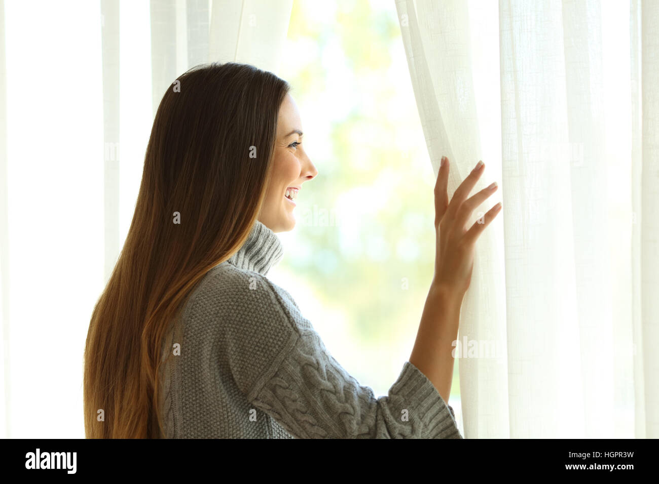 Vista laterale di una donna felice di aprire le tende di una finestra e godendo di un nuovo giorno con una calda luce da esterno Foto Stock