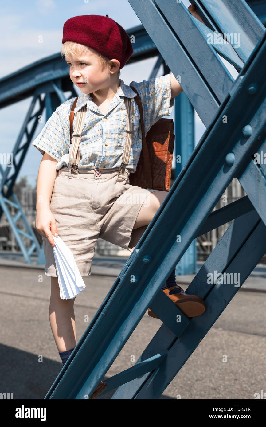 In stile retrò carino boy presso la strada - dalla scuola indossare sacchetta, autoreggenti, magliette, pantaloncini corti e berretto rosso hat, giocare con aeroplano di carta e di salire al blu Foto Stock