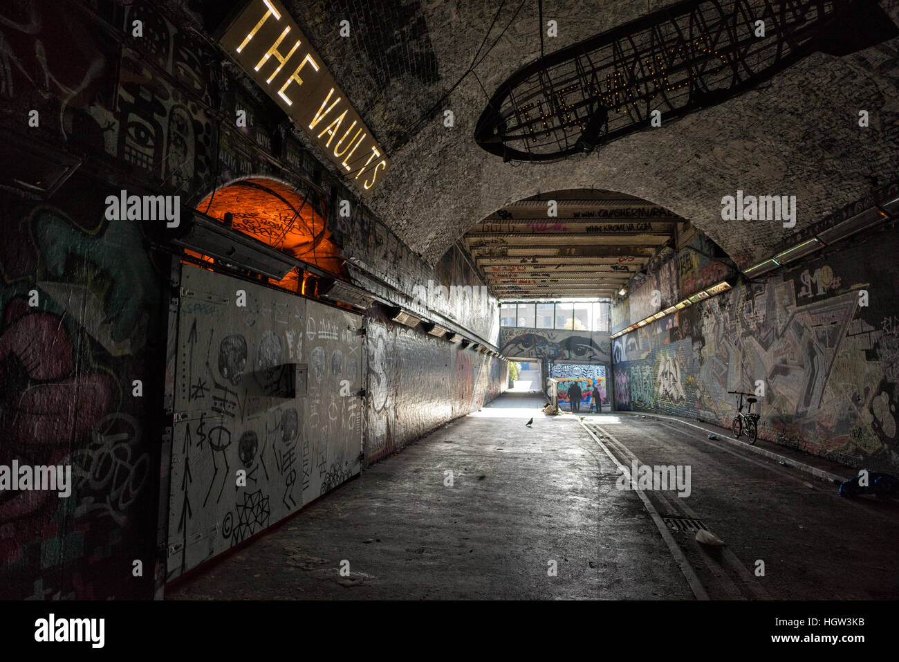 Tunnel in mattoni ricoperti di graffiti in Leake Street, Londra, con il cartello illuminato "The Vaults" e la luce del giorno che si riversa attraverso l'estremità della strada urbana Foto Stock