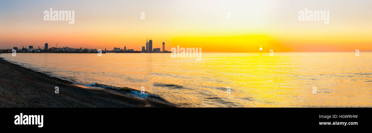 Batumi, Adjara, Georgia. Panorama della città di villeggiatura al tramonto Sunrise. Luminose del cielo della sera in colori di giallo e arancione. Vista dal mare spiaggia a Cityscape W Foto Stock