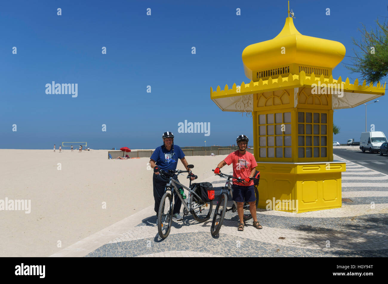 Due persone senior in marcia del ciclo con le biciclette da un giallo beach kiosk presso la spiaggia di Foz do Arelho sulla Costa d'argento del Portogallo Foto Stock