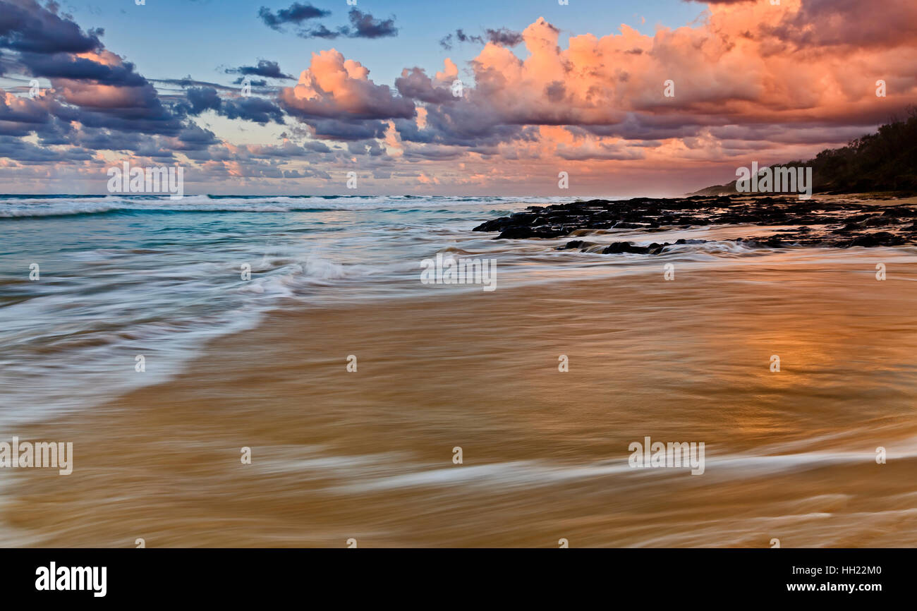 Seascape sunrise oltre 75 miglia di spiaggia su Fraser Island nel Queensland, in Australia. Vulcaniche nere pietre di granito stand contro pacific surf e maree a Foto Stock