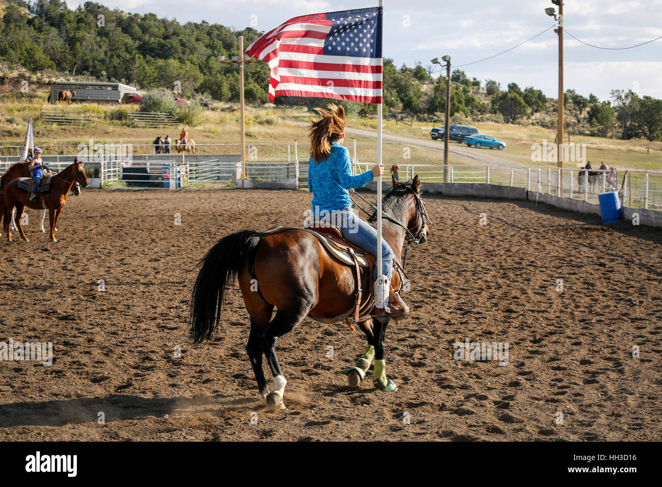 Giovane donna in sella il suo cavallo attraverso il rodeo arena portando la bandiera americana durante la stella lamas banner in una gioventù rodeo Foto Stock