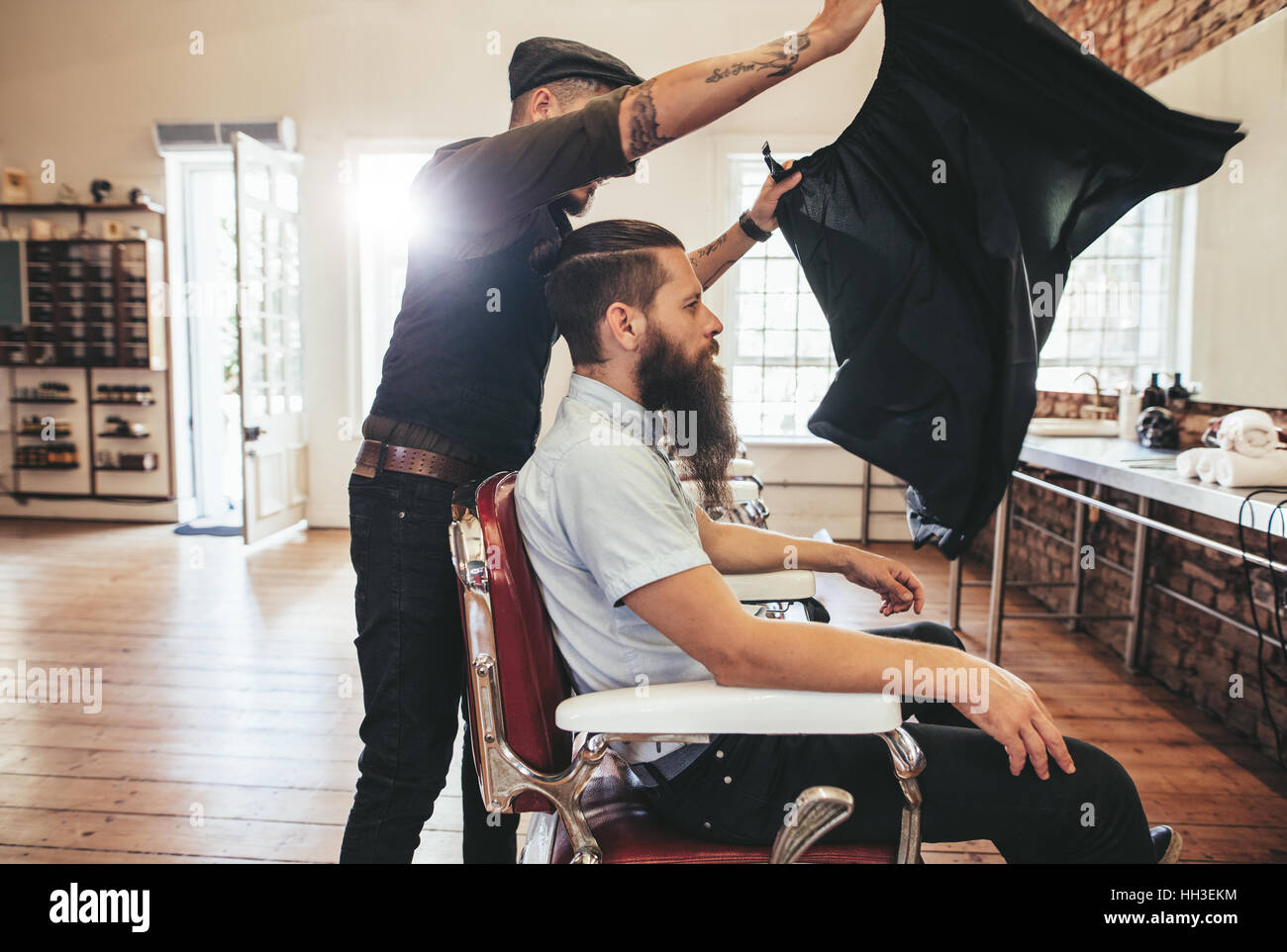 Barbiere di inserire un foglio per coprire il suo cliente da tagliare i capelli. Parrucchiere maschio al lavoro con il bel uomo seduto sulla sedia. Foto Stock