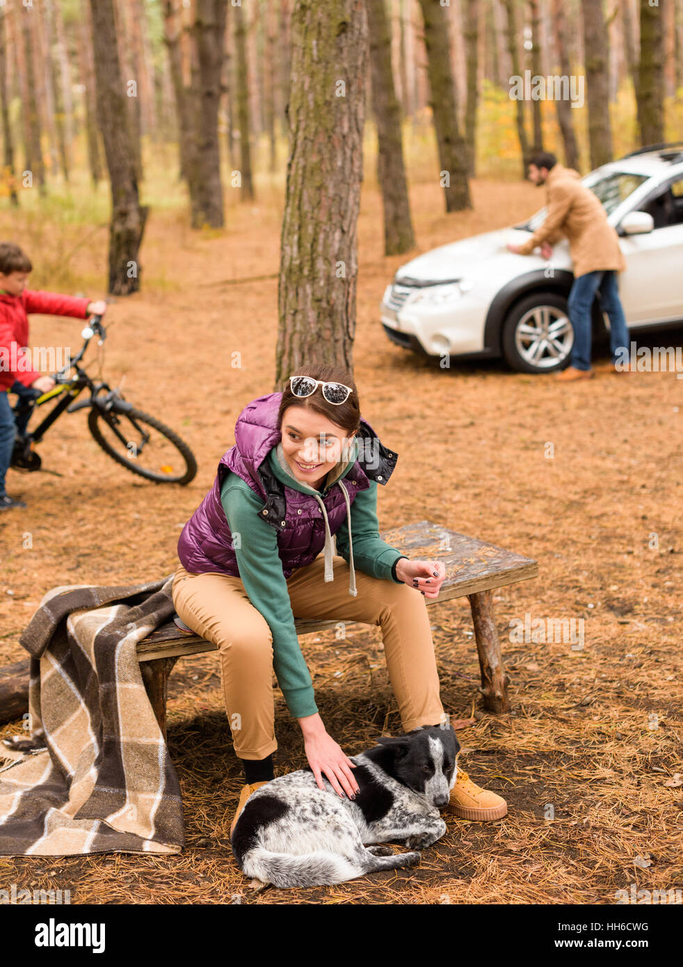 Donna sorridente seduta sul banco e accarezzare un cane in autunno foresta con uomo sfocata auto pulizia e ragazzo Bicicletta Equitazione su sfondo Foto Stock