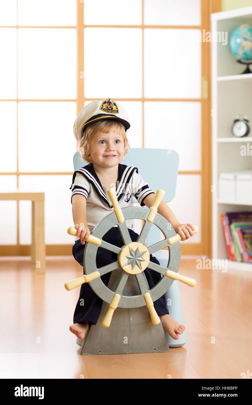 Bambino vestito come un capitano o un marinaio gioca sulla sedia come a bordo di una nave nella sua stanza. Bambino ragazzo ruota il volante in legno. Foto Stock