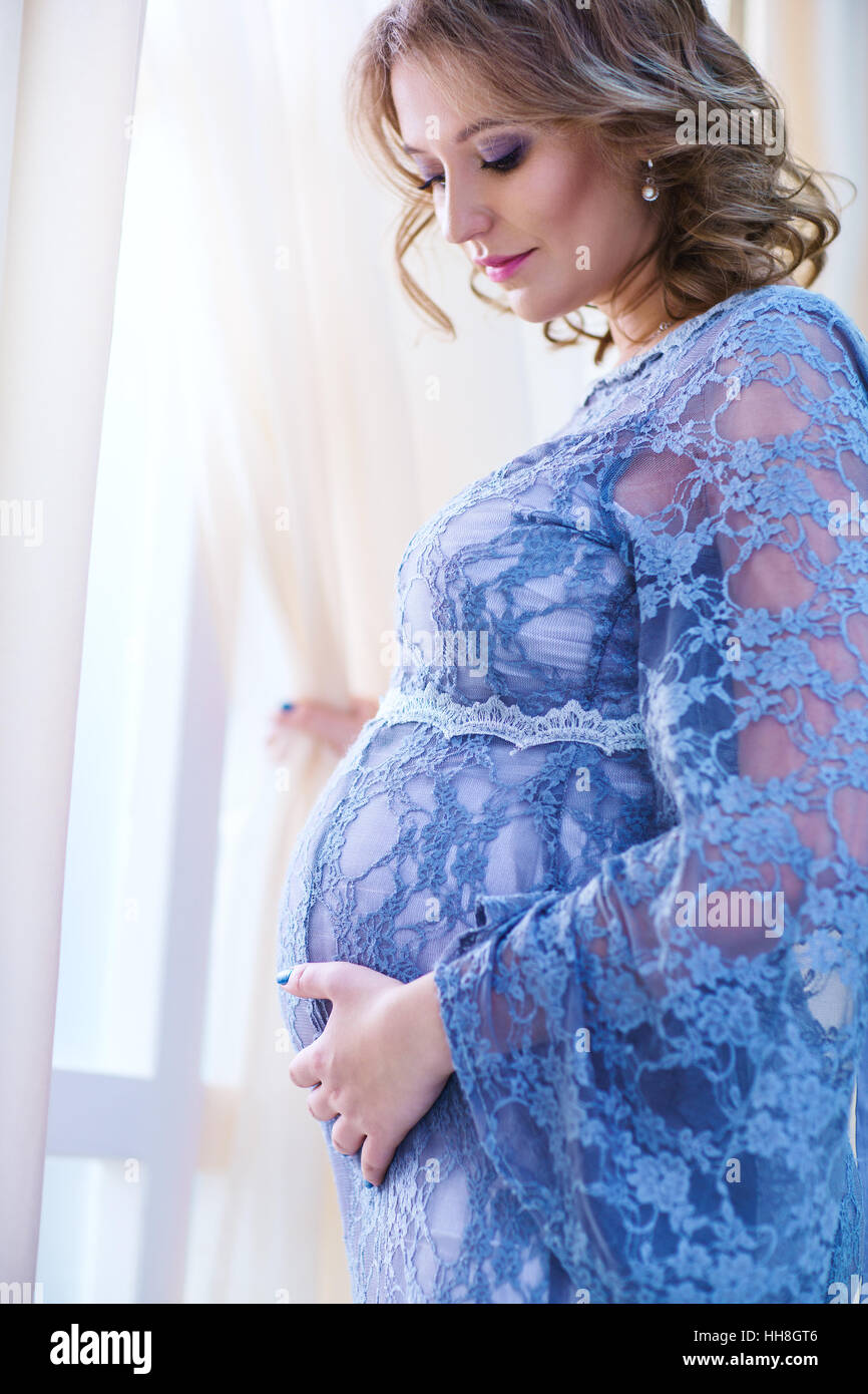 Giovane donna incinta in piedi alla finestra ritratto in interni in colori tenui Foto Stock