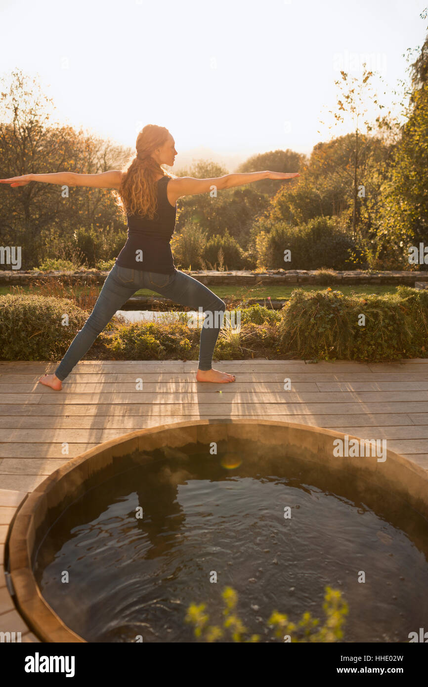 La donna a praticare yoga warrior 2 pone in autunno di un patio con vasca idromassaggio Foto Stock