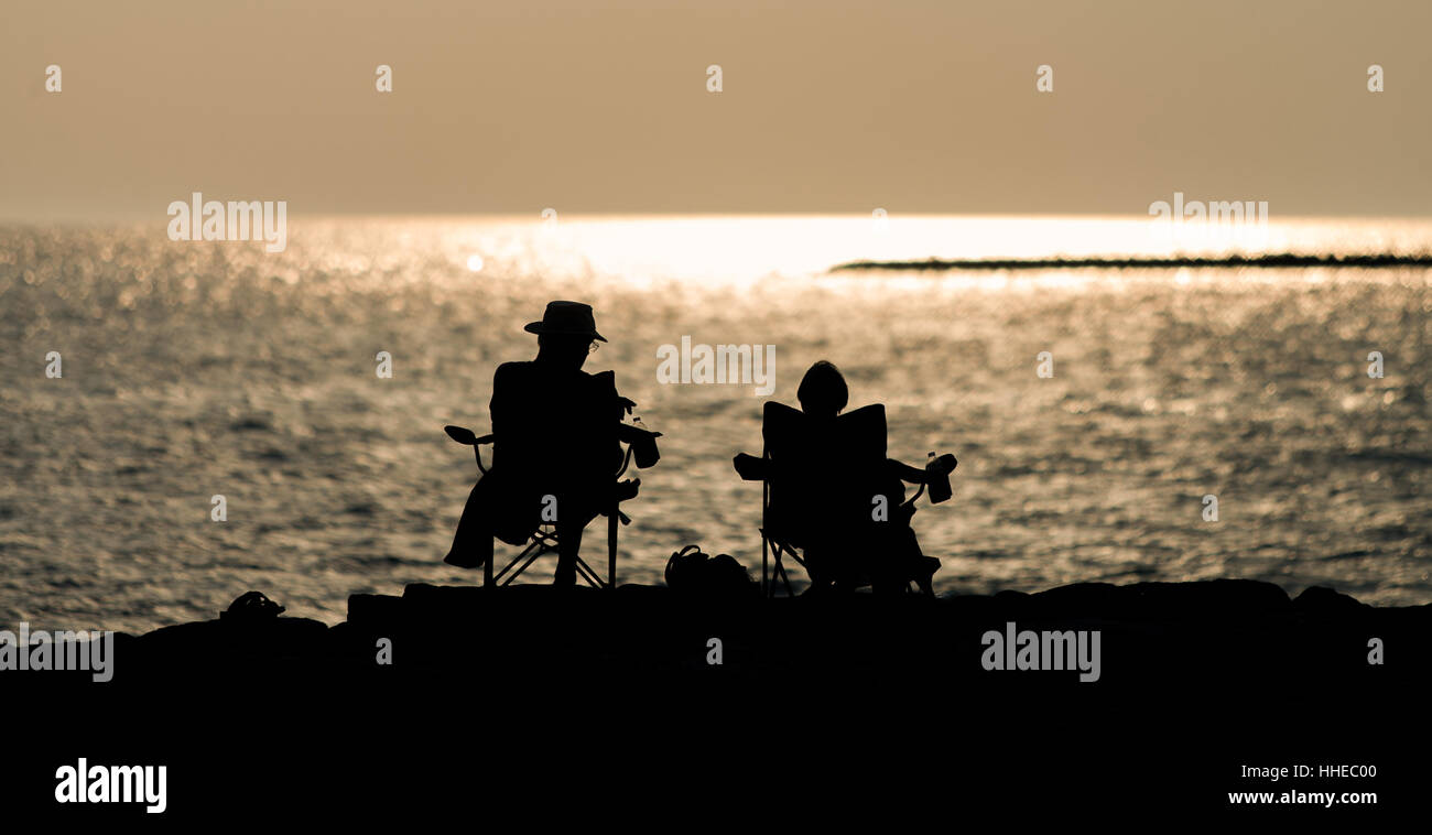 Un uomo e una donna seduti nelle sedie lungo il litorale del Lago Huron e godetevi il tramonto come uccelli e barche dal galleggiante. Foto Stock