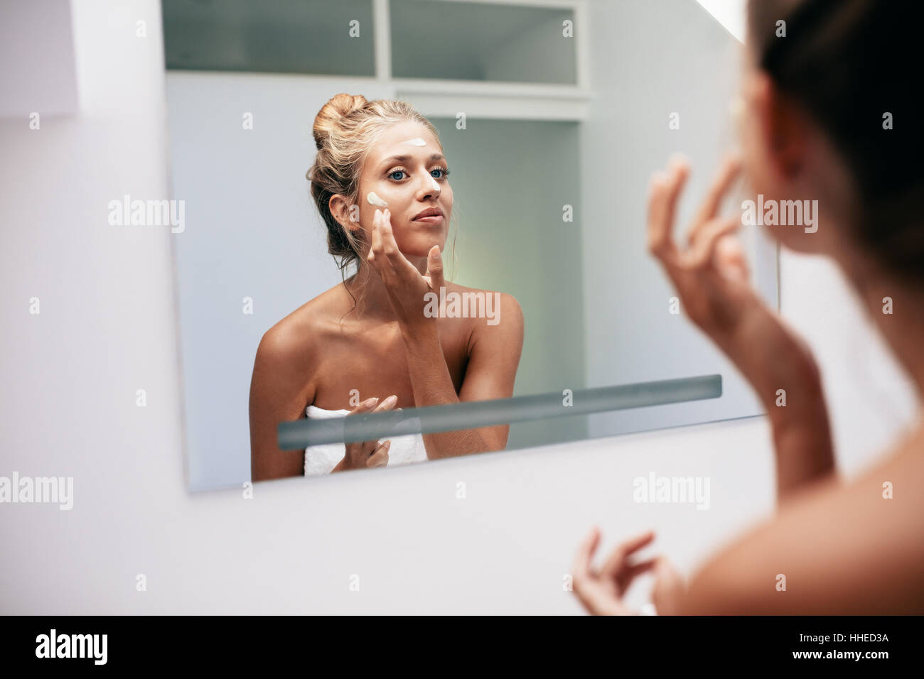Giovane donna di riflessione a specchio in applicazione di crema nel bagno. Femmina caucasica facendo un trattamento di bellezza sul suo viso a casa. Foto Stock