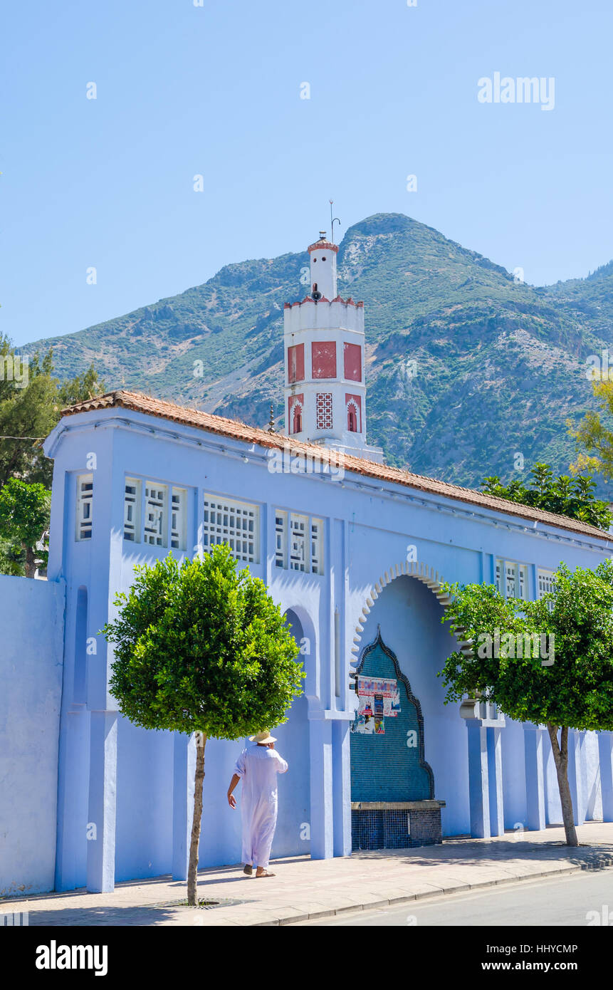 Uomo arabo in piedi di fronte blu lavato a parete, moschea rossa e la montagna in città Chefchaouen, Marocco, Africa del Nord. Foto Stock