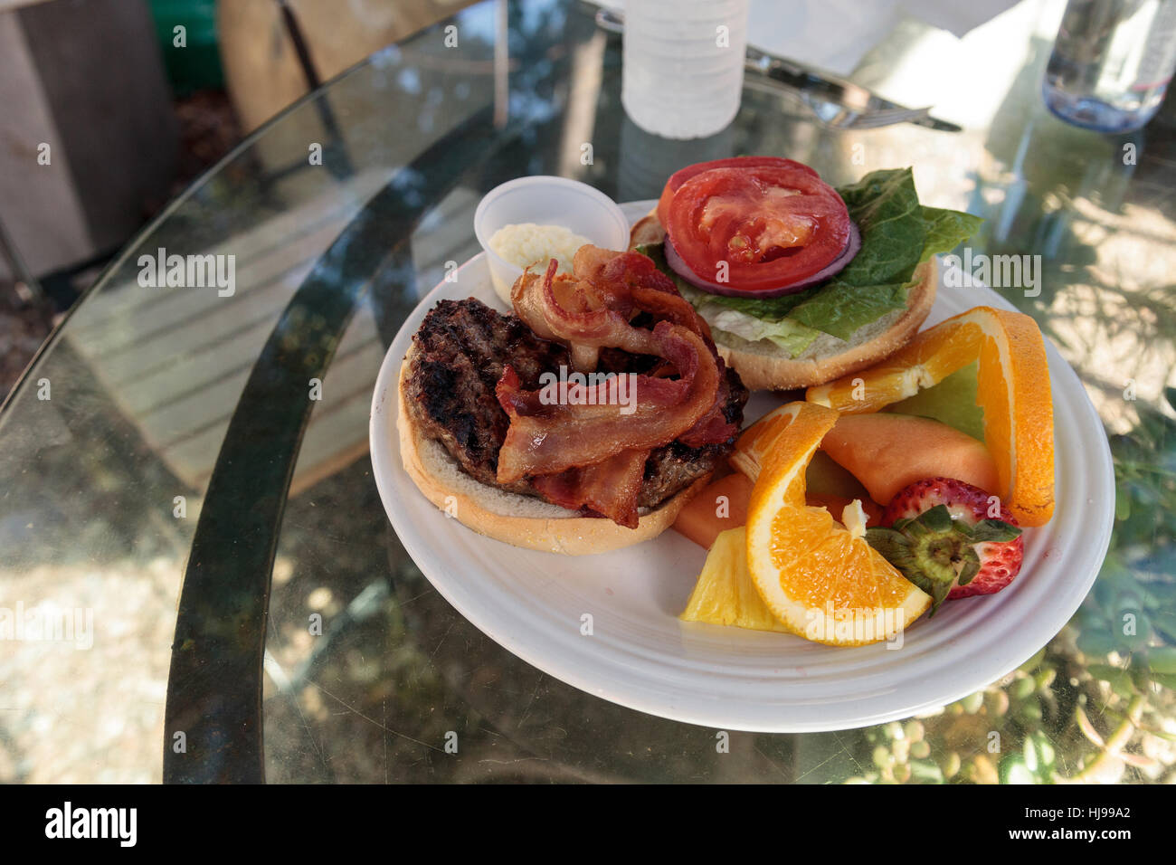 Hamburger con pancetta sulla parte superiore su un soffice focaccia di pane e di un lato della frutta comprese le arance, fragole, meloni e ananassi per l Foto Stock